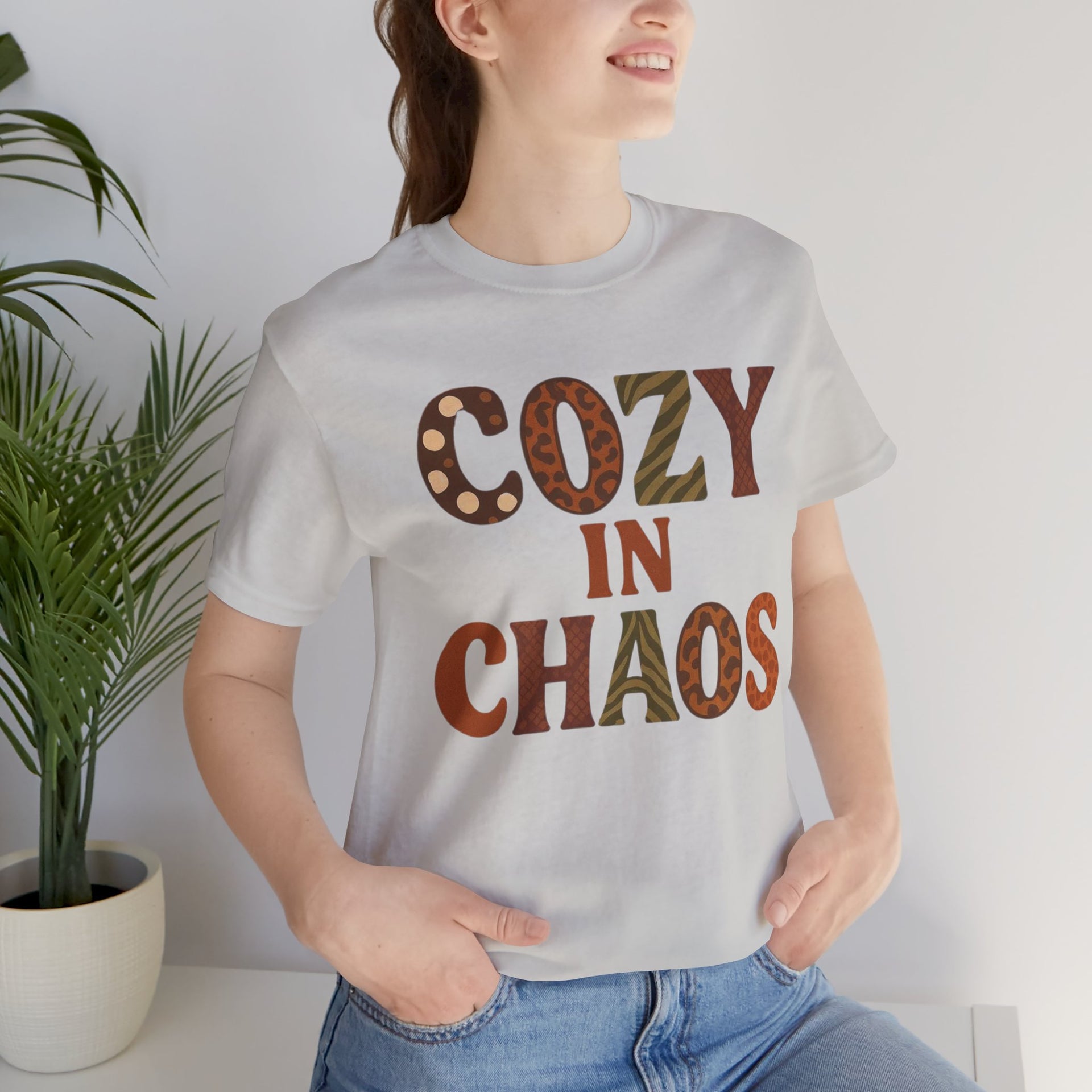 Person wearing a white t-shirt with 'Cozy in Chaos' text, standing indoors with a plant in the background.