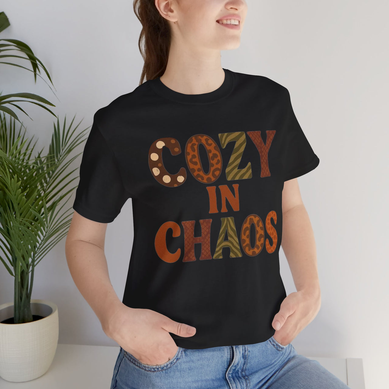 Person wearing a black t-shirt with 'Cozy in Chaos' text, standing indoors next to a plant.