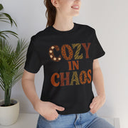 Person wearing a black t-shirt with 'Cozy in Chaos' text, standing indoors next to a plant.