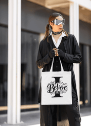 Person holding a tote bag with 'I Believe' design outdoors.