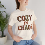 Person wearing a white t-shirt with 'COZY IN CHAOS' text, standing indoors next to a plant.