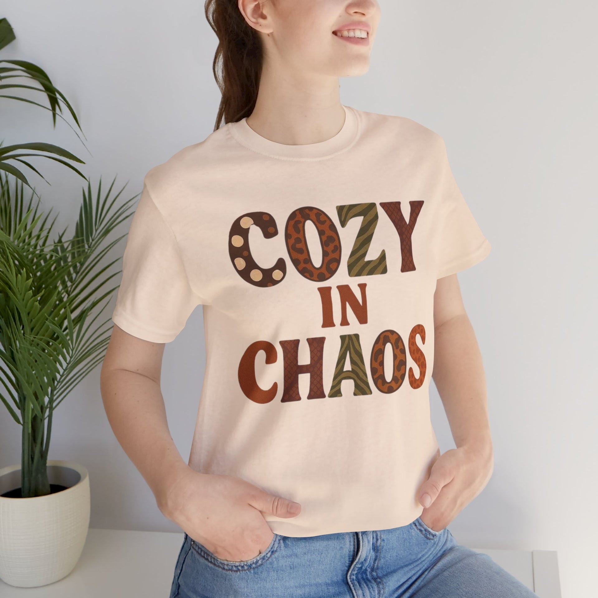 Person wearing a beige t-shirt with 'Cozy in Chaos' text, standing indoors next to a plant.