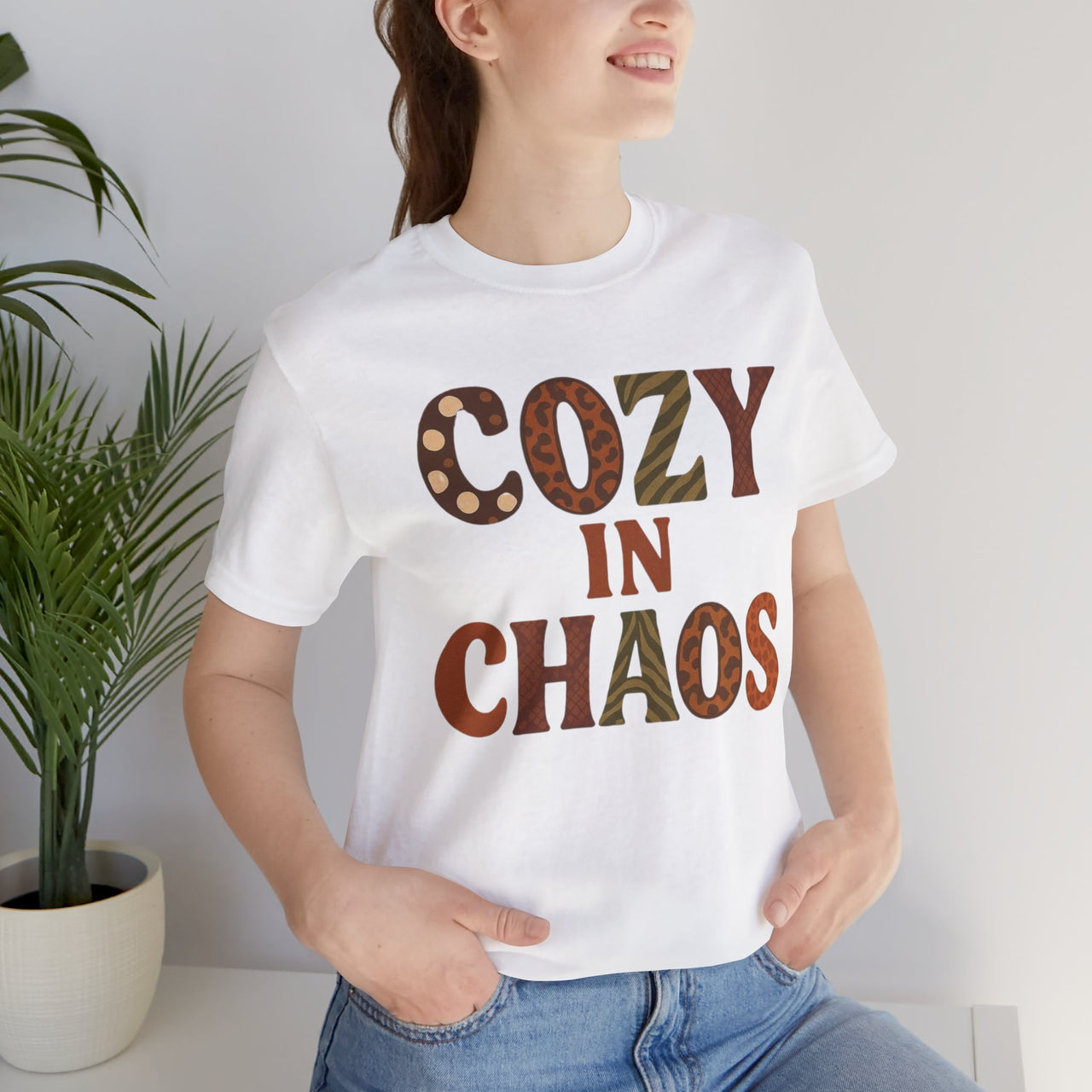 Person wearing a white t-shirt with 'Cozy in Chaos' text, standing indoors next to a plant.