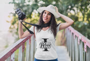 Woman wearing a white t-shirt with 'I Believe' design, holding a camera on a bridge.