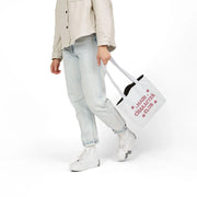 Person holding a white tote bag with text, wearing light blue jeans and a beige jacket on a white background