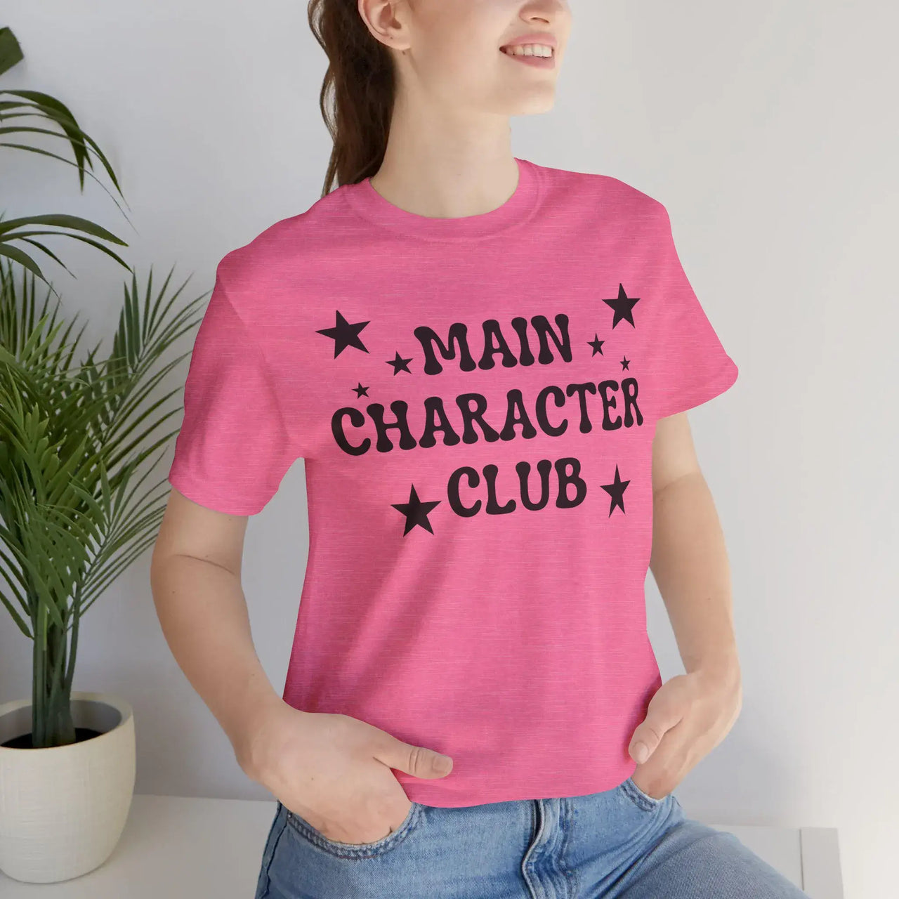 Person wearing a pink t-shirt with 'MAIN CHARACTER CLUB' text and stars, standing against a white wall with a plant on the left.
