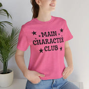 Person wearing a pink t-shirt with 'MAIN CHARACTER CLUB' text and stars, standing against a white wall with a plant on the left.