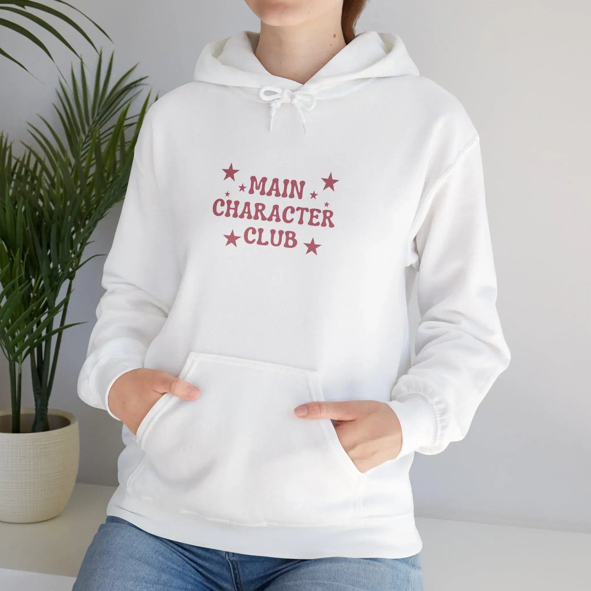 White hoodie with 'Main Character Club' text and stars worn by a person, against a neutral background.
