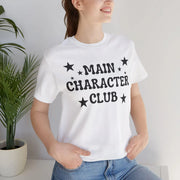 Person wearing a white t-shirt with 'MAIN CHARACTER CLUB' text and stars, standing against a plain background.