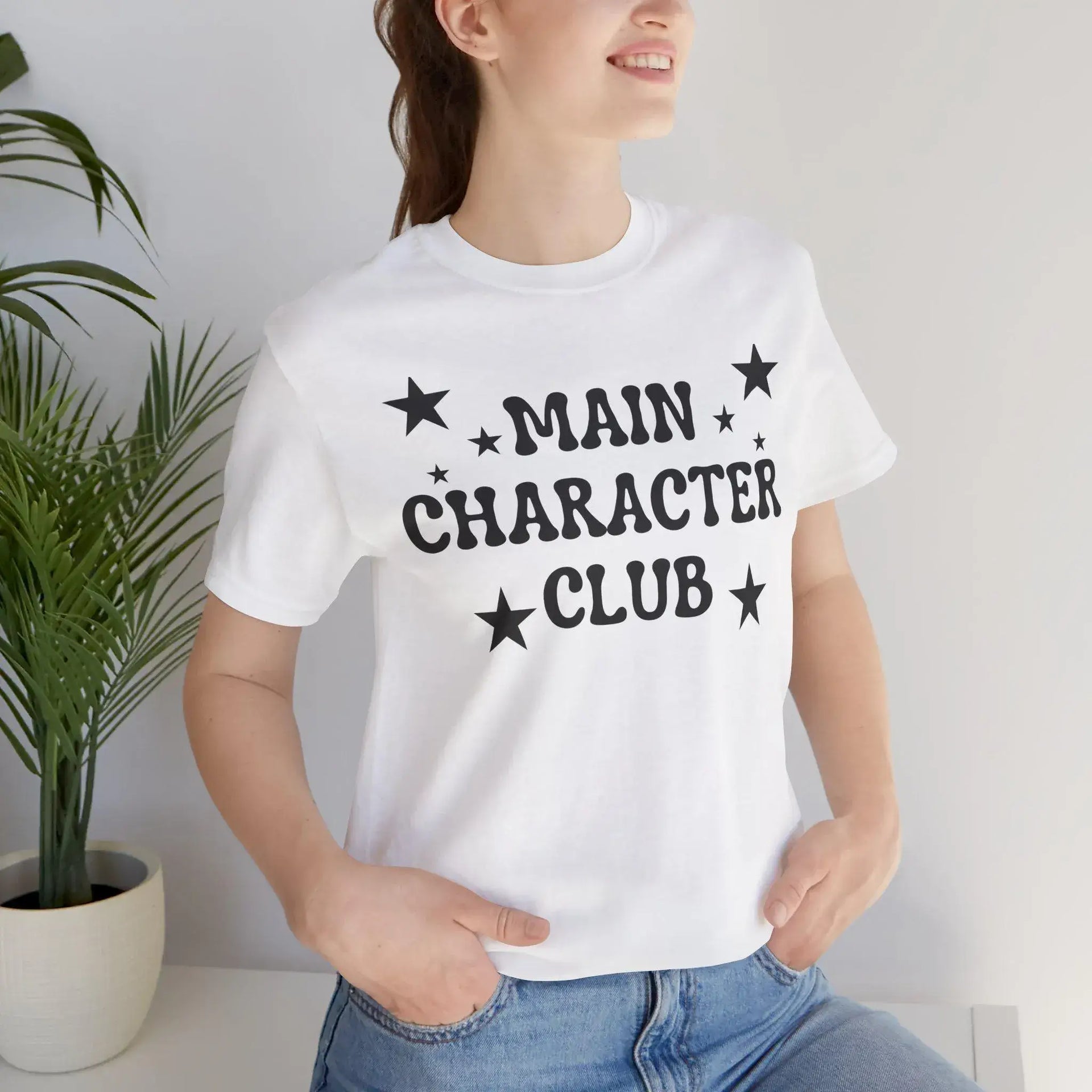 Person wearing a white t-shirt with 'MAIN CHARACTER CLUB' text and stars, standing against a plain background.