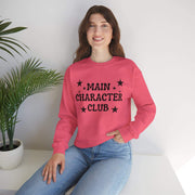 Person wearing a pink sweatshirt with 'MAIN CHARACTER CLUB' text, sitting against a white wall with a plant.