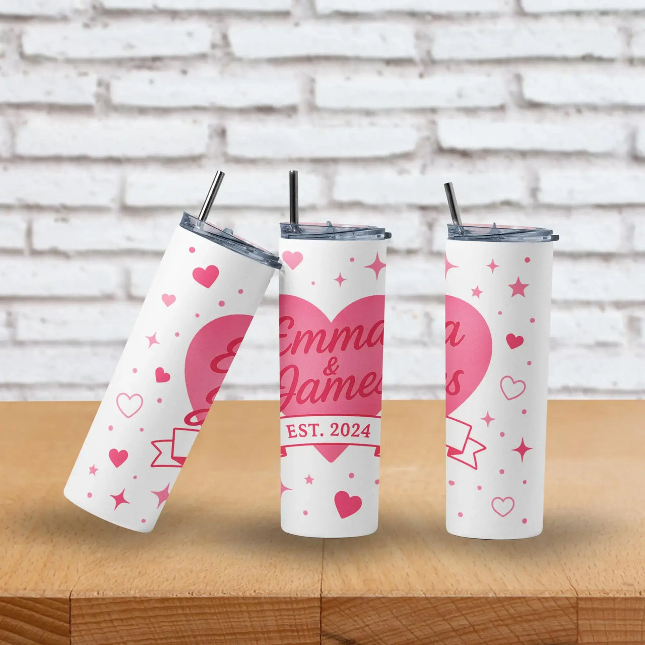 Three personalized tumblers with hearts and text on a wooden surface and brick wall background.