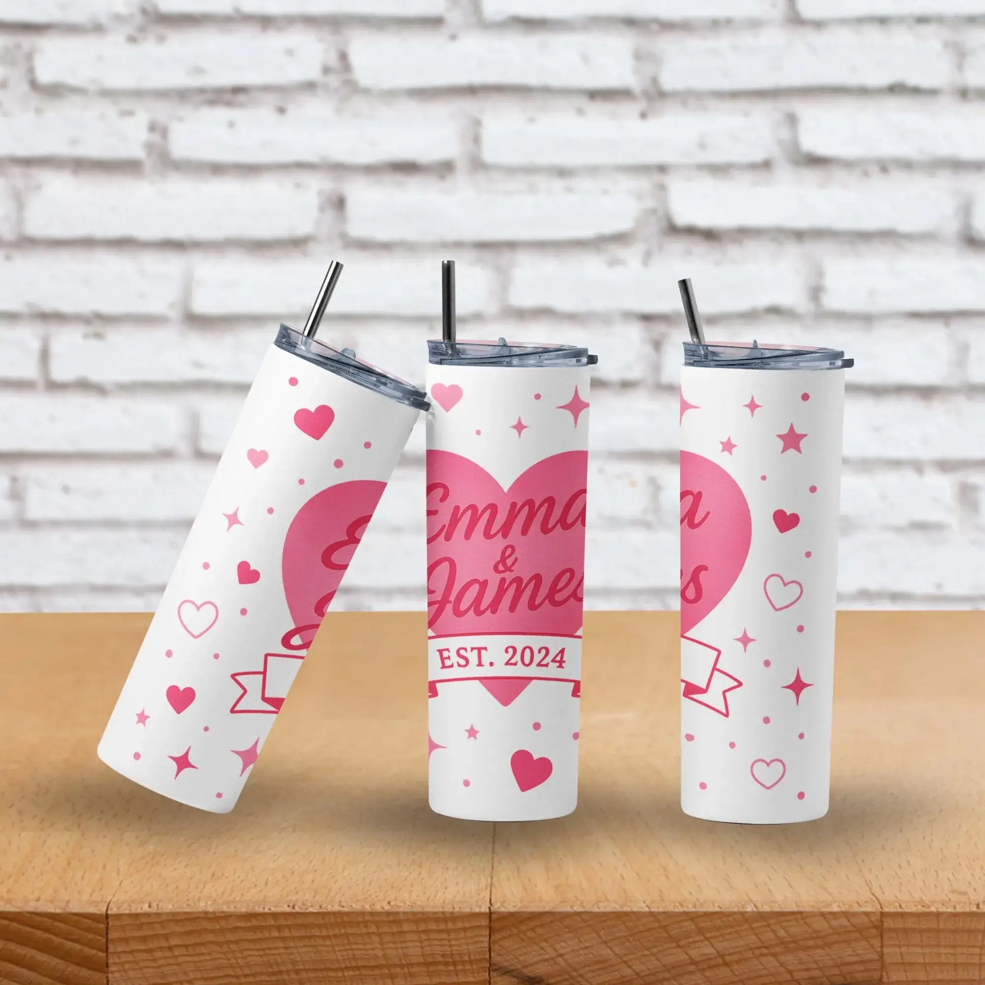 Three personalized tumblers with hearts and text on a wooden surface and brick wall background.