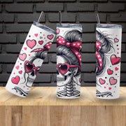 Three insulated tumblers with skull designs, hearts, and bows on a wooden surface with a brick wall background.