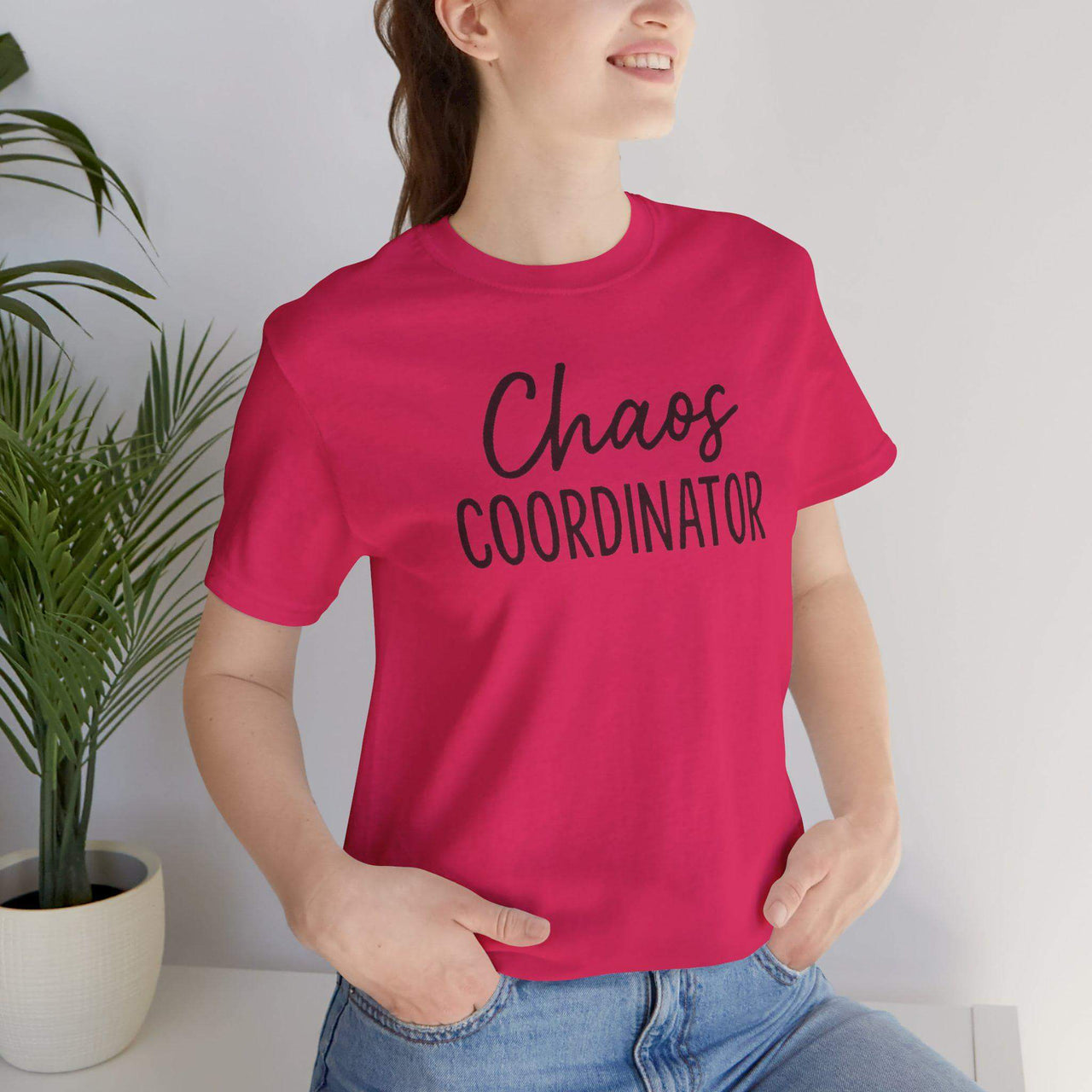 Person wearing a pink t-shirt with 'Chaos Coordinator' text, standing indoors next to a plant.