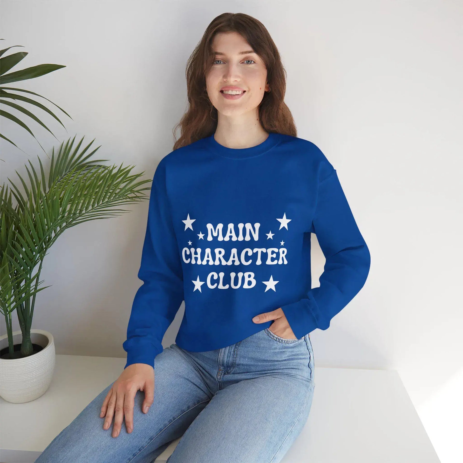 Person wearing a blue sweatshirt with 'MAIN CHARACTER CLUB' text, sitting indoors.