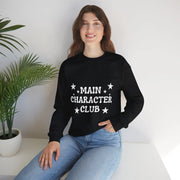 Person wearing a black sweatshirt with 'MAIN CHARACTER CLUB' text, sitting on a white surface with a plant in the background.