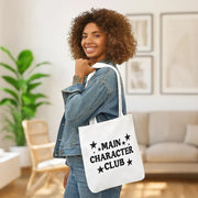 Woman holding a tote bag with 'Main Character Club' text in a living room setting