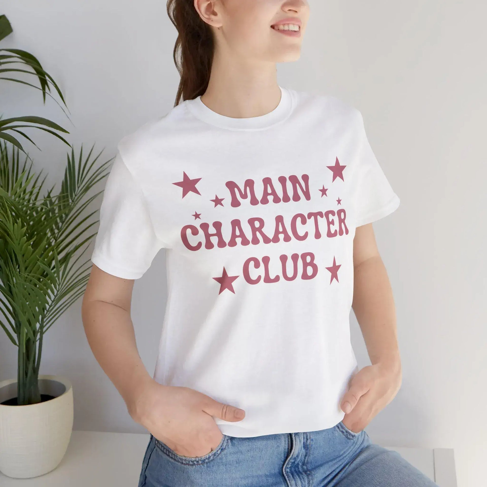 Person wearing a white t-shirt with 'MAIN CHARACTER CLUB' text and stars, standing indoors.