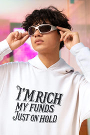 Person wearing a white hoodie with text 'I'm rich, my funds just on hold' against a blurred background.