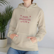 Beige hoodie with 'Main Character Club' text and stars, worn by a person, against a plain background.