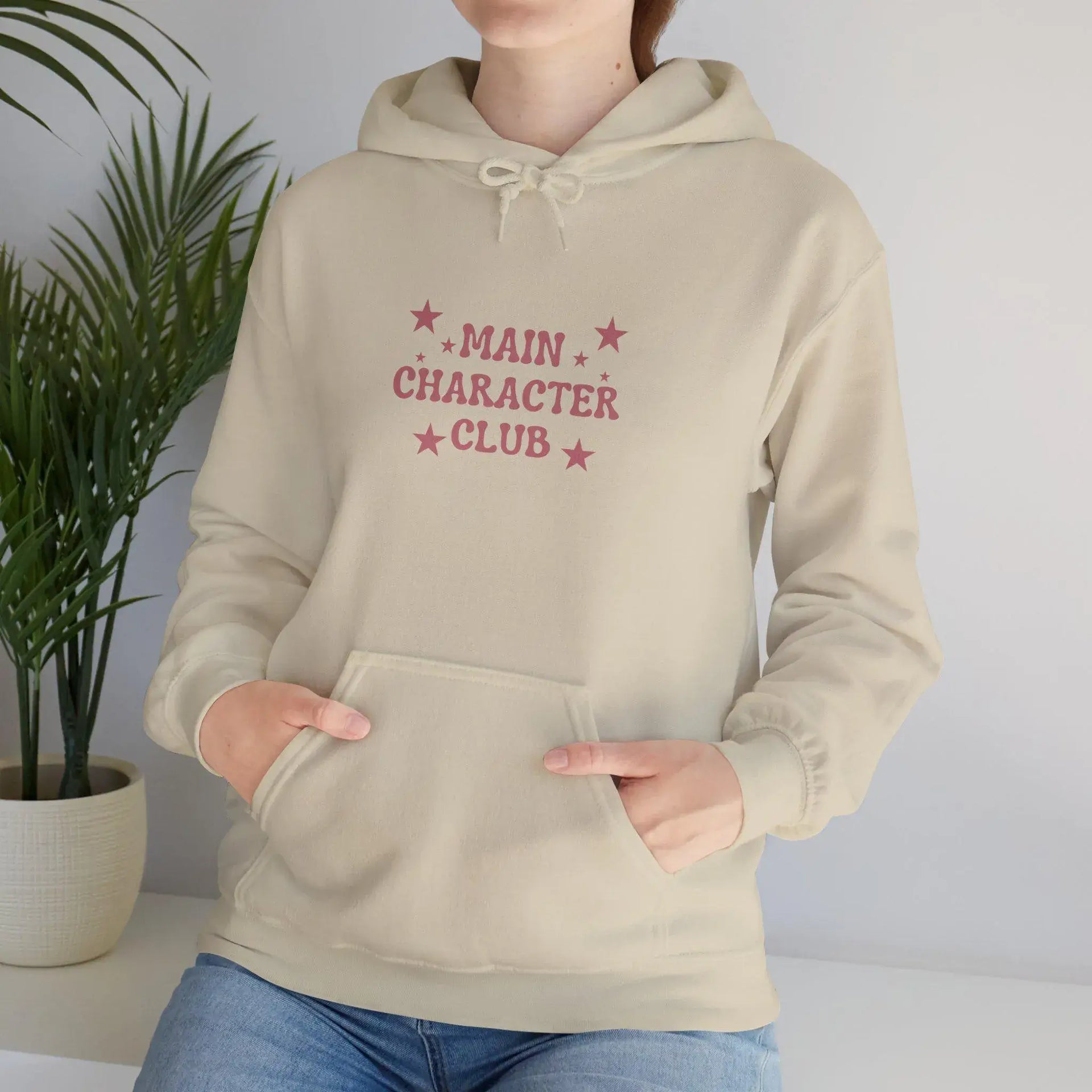 Beige hoodie with 'Main Character Club' text and stars, worn by a person, against a plain background.