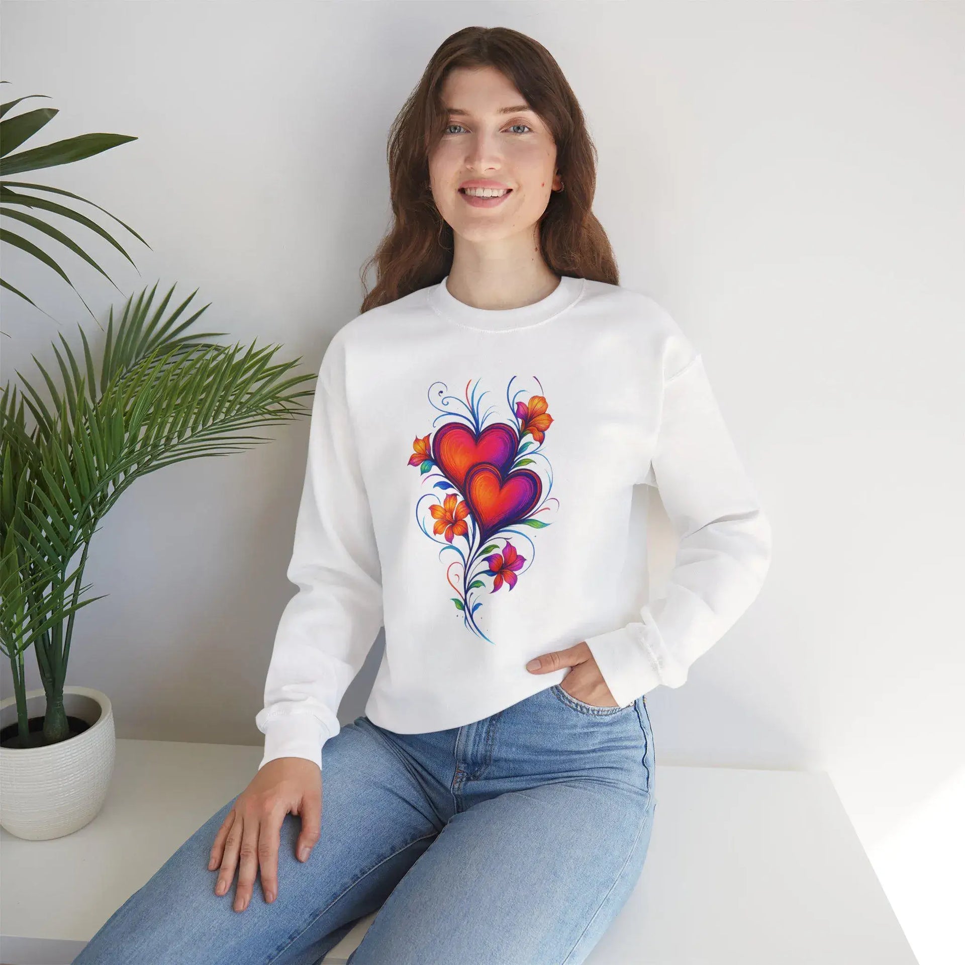 Woman wearing a white sweatshirt with a colorful heart design, sitting in a room with a plant.