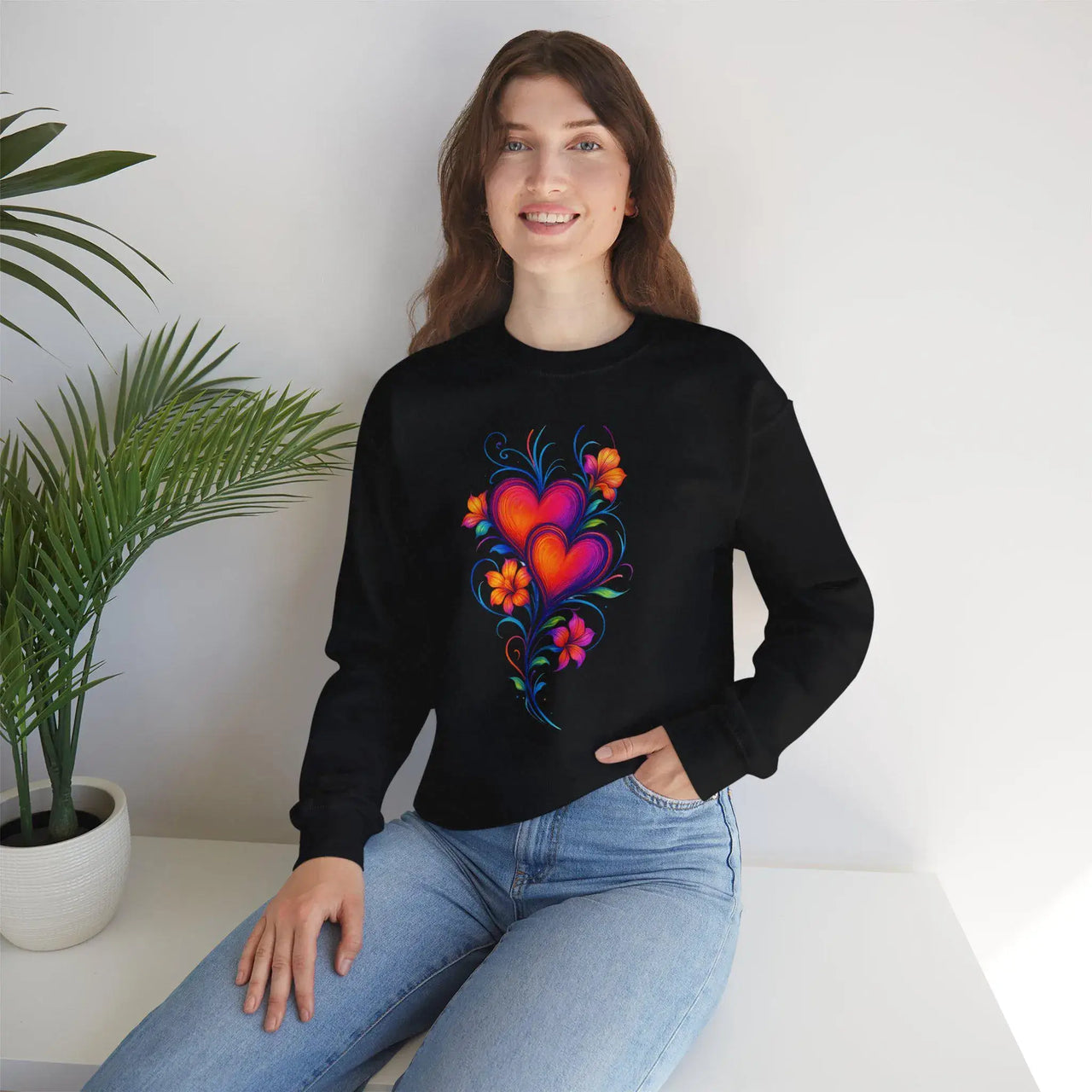 Person wearing a black sweatshirt with a colorful heart design, sitting on a white surface with a plant in the background.