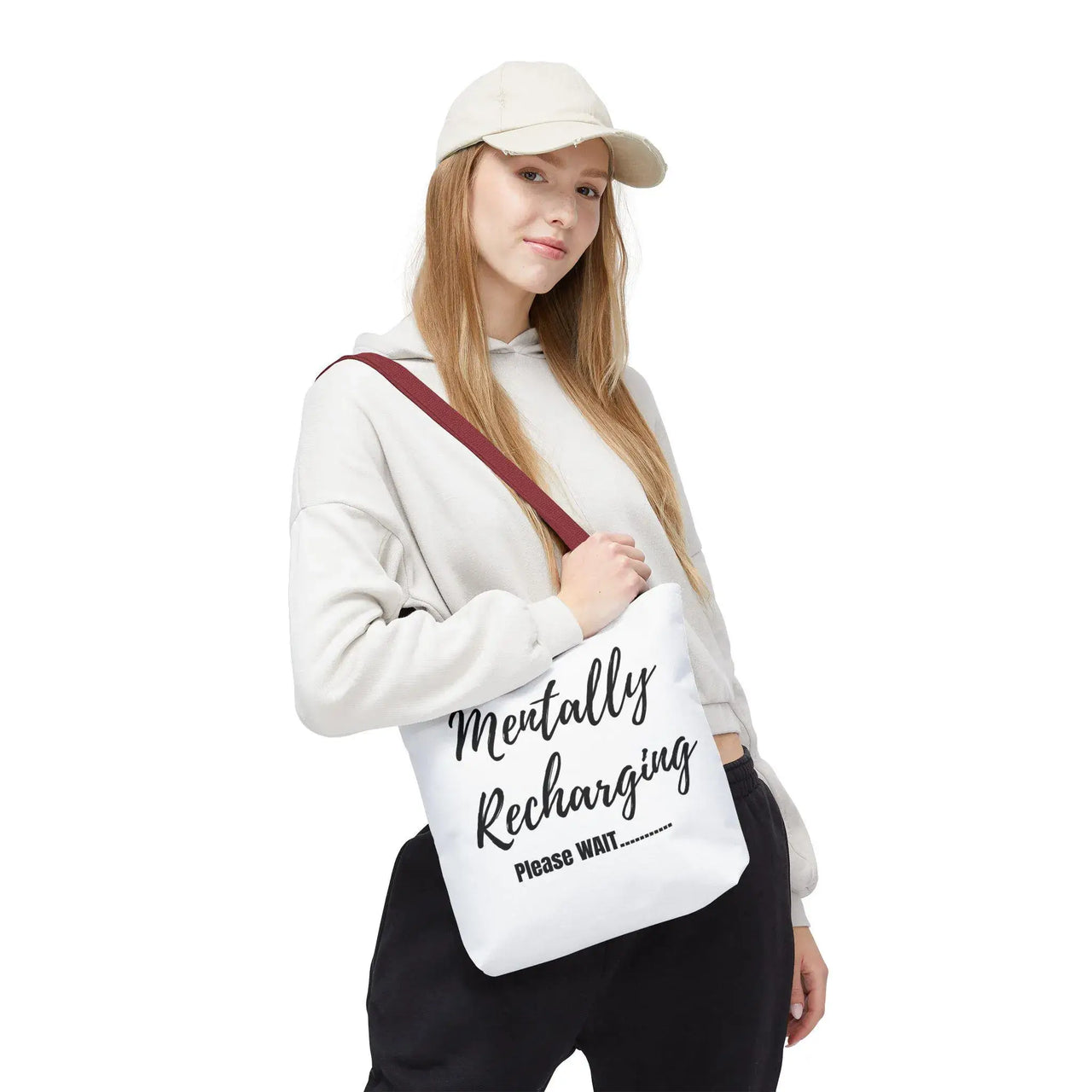 Person holding a white tote bag with text, wearing a cap and white hoodie on a white background