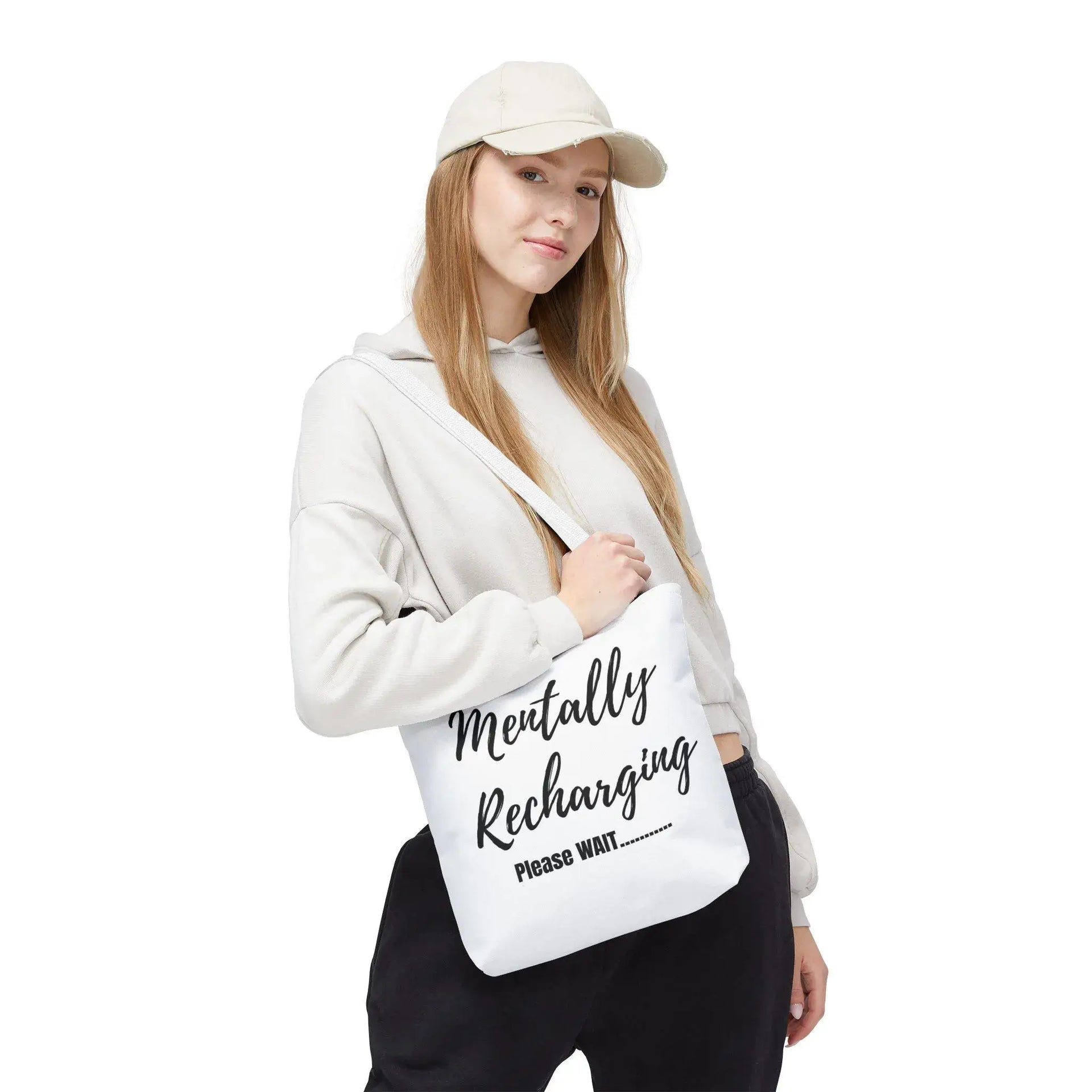 Person holding a white tote bag with text, wearing a white hoodie and cap on a white background