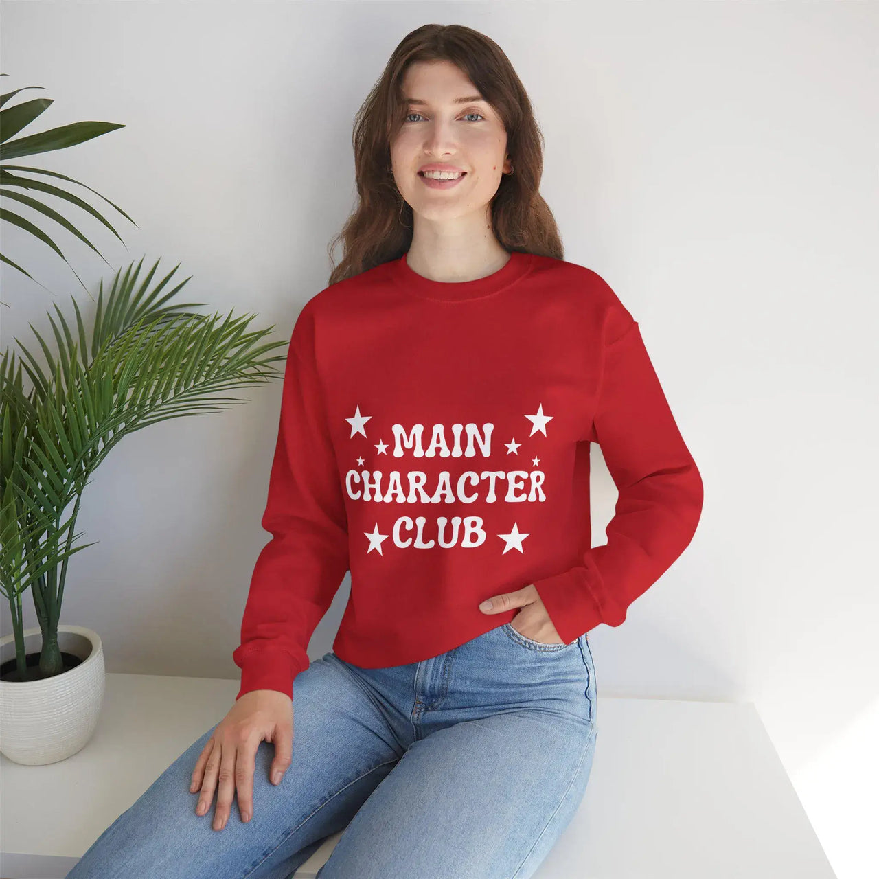 Person wearing a red sweatshirt with 'MAIN CHARACTÓR CLUB' text, sitting on a white surface with a plant in the background.