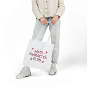 Person holding a white tote bag with 'Main Character Club' text and star designs.