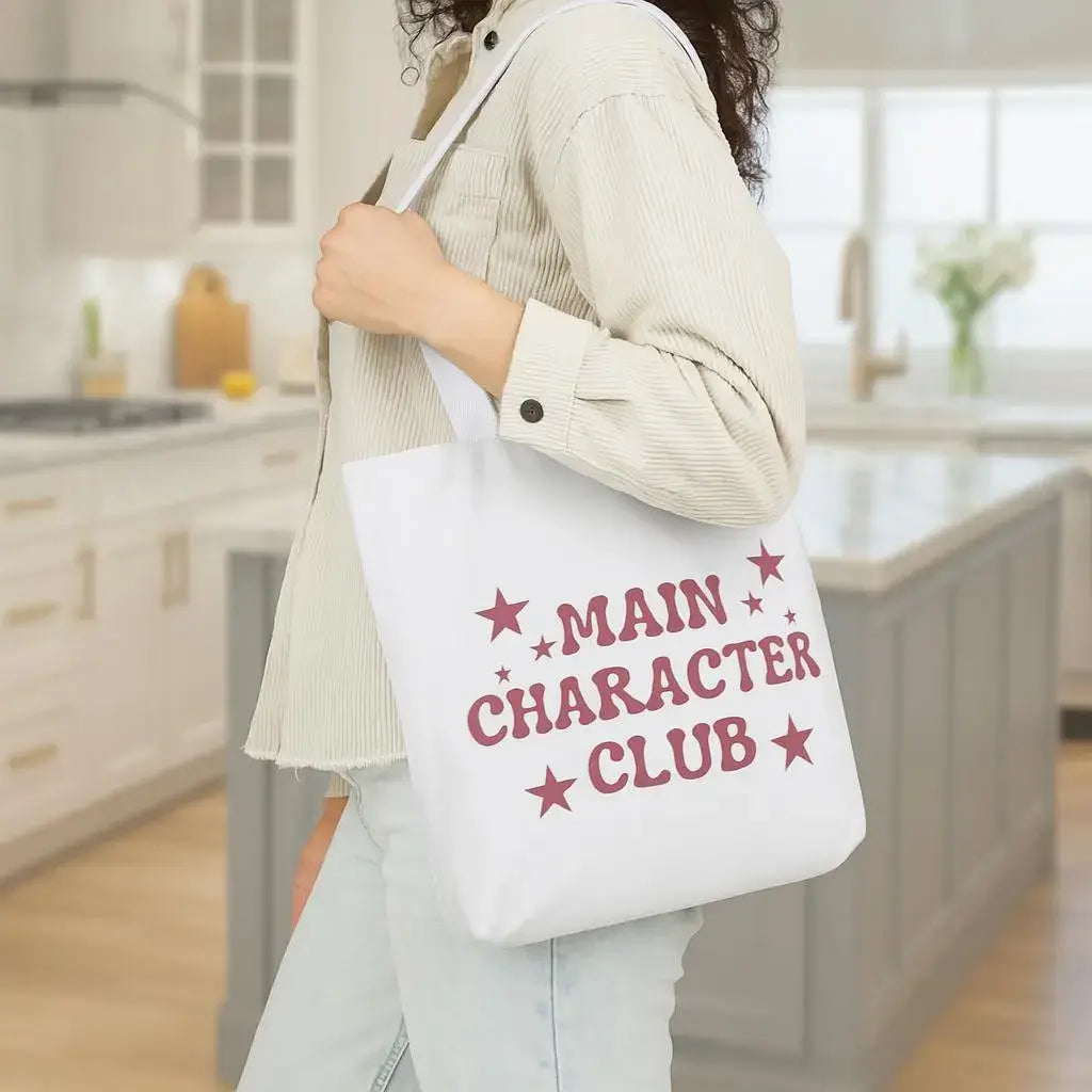 Person holding a tote bag with 'MAIN CHARACTER CLUB' text in a kitchen setting