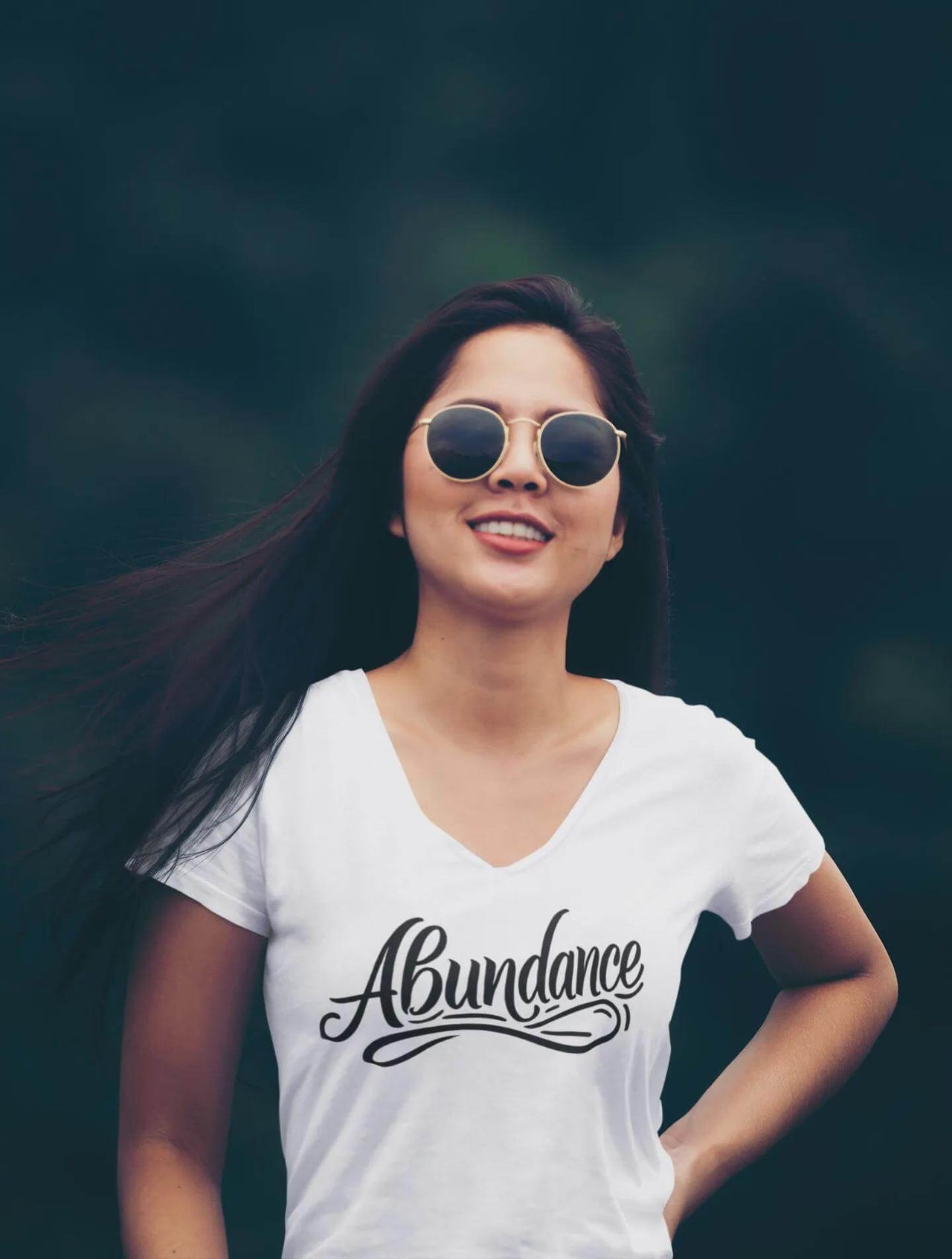 Woman wearing a white t-shirt with 'Abundance' text, standing against a dark background.