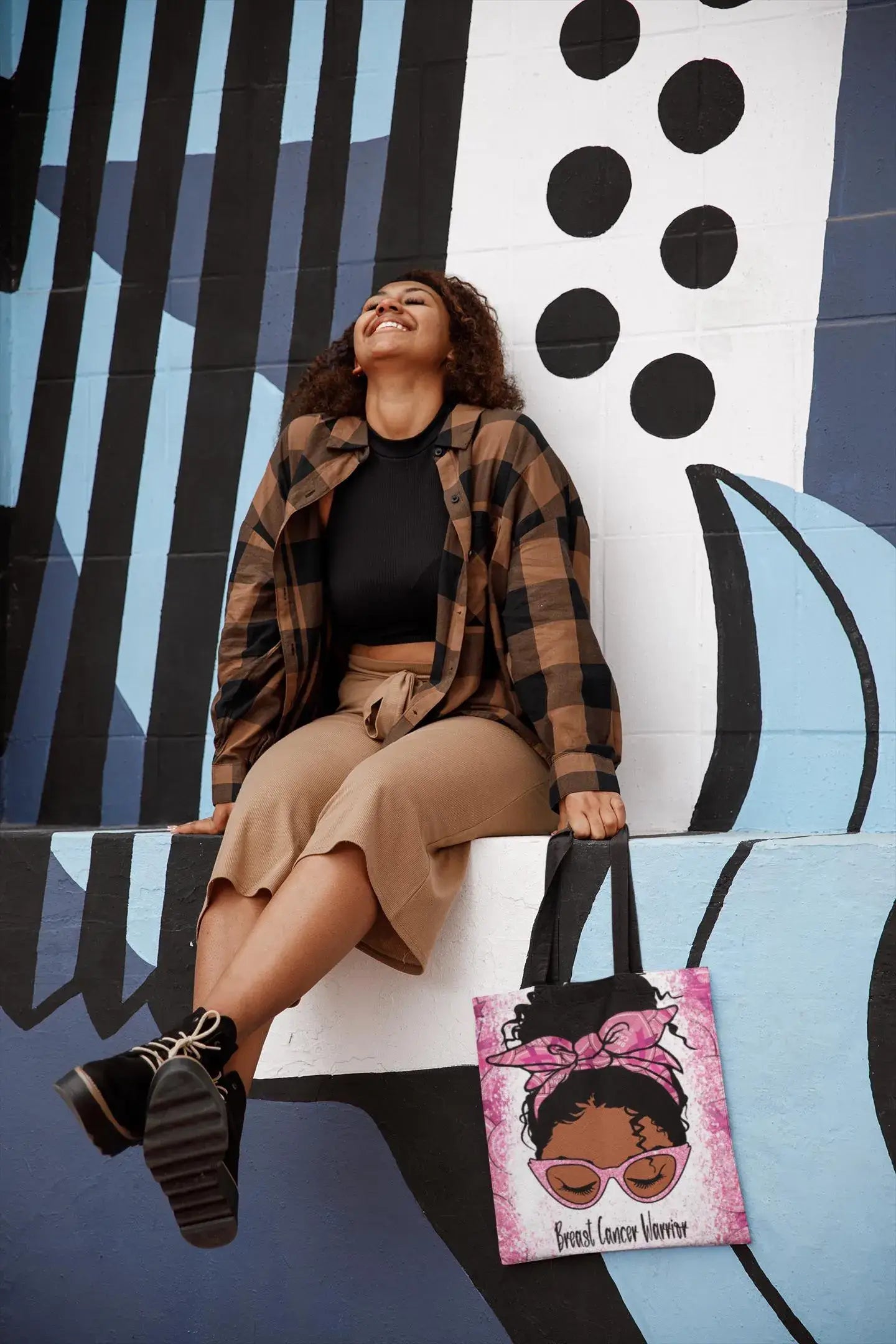 Woman sitting on a wall with a colorful abstract design, holding a tote bag with an illustration of a woman with a pink bow.