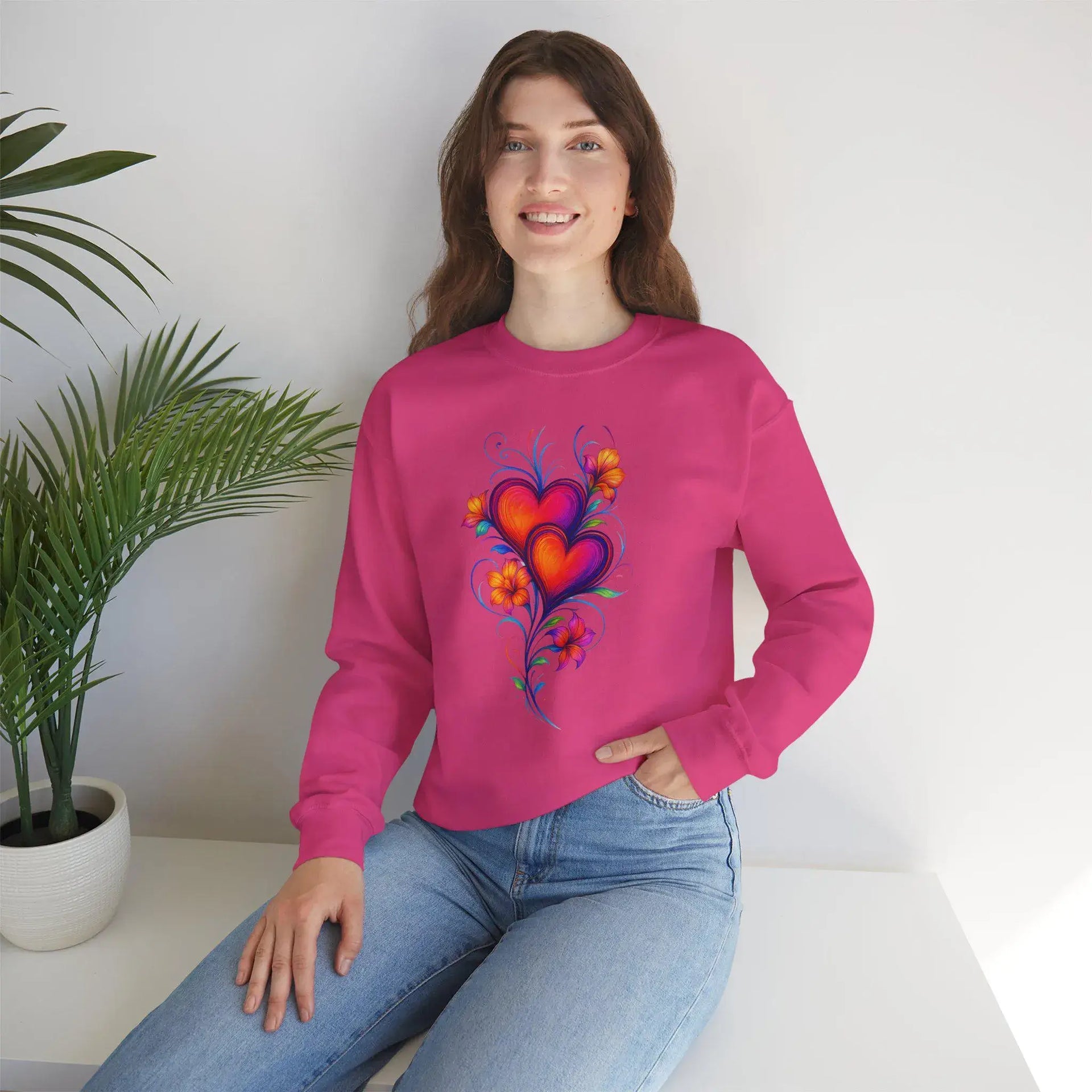 Person wearing a pink sweatshirt with a colorful heart design, sitting against a white wall with a plant.