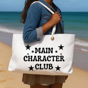 Person holding a tote bag with 'Main Character Club' text on a beach