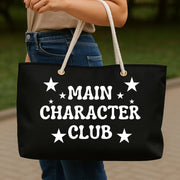 Black tote bag with 'MAIN CHARACTER CLUB' text held by a person outdoors.