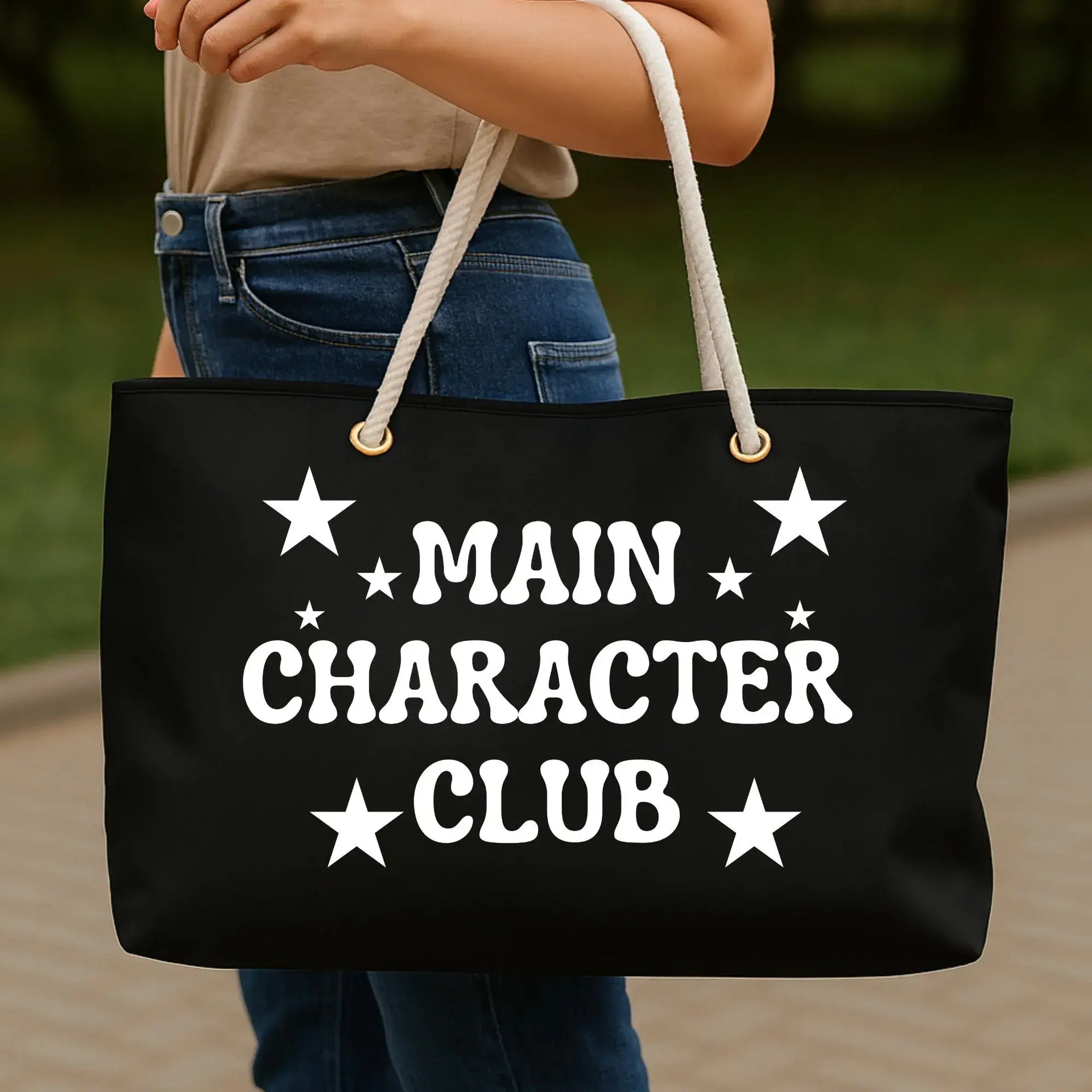 Black tote bag with 'MAIN CHARACTER CLUB' text held by a person outdoors.