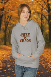 Person wearing a gray hoodie with 'Cozy in Chaos' text in an autumn setting
